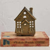 A bronze-colored embossed cast aluminum house tealight holder with open panes and a pointed roof line, placed on top of a cardboard box with 'Perfecto Grande' printed on it.