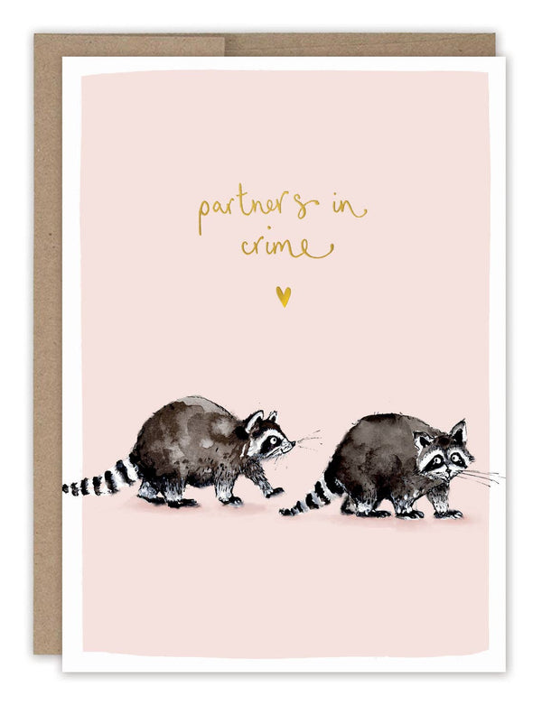 A greeting card with an illustration of two raccoons and the text 'partners in crime' on a pink background. The card is accompanied by a brown Kraft envelope.