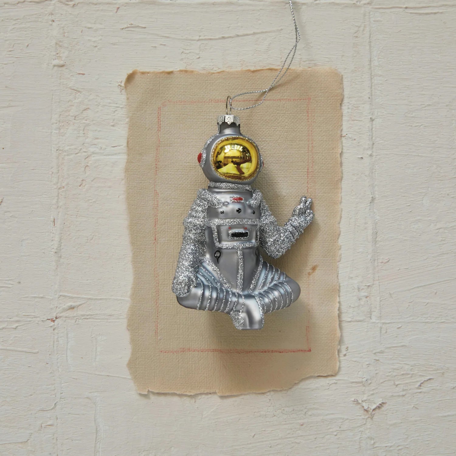 A hand-painted glass astronaut ornament with a peace sign and glitter, featuring a silver and gold finish, displayed on a textured wall.