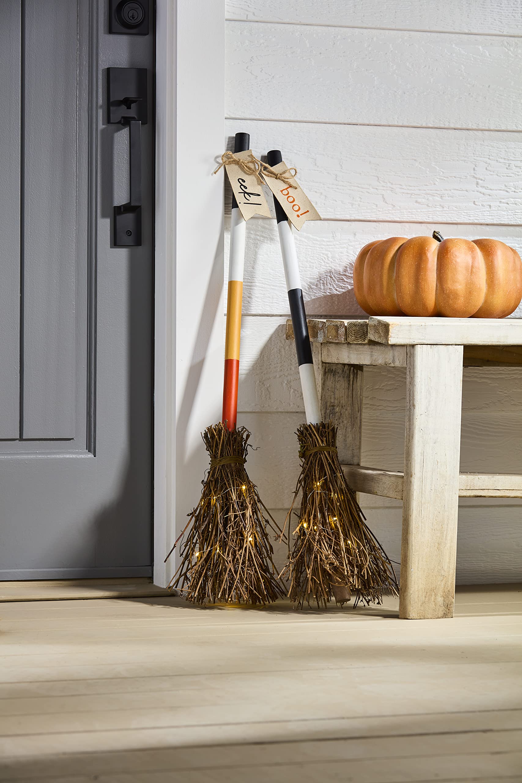 Two brooms with lights on a wooden stool next to a pumpkin against a white door.