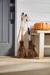 Two brooms with lights on a wooden stool next to a pumpkin against a white door.