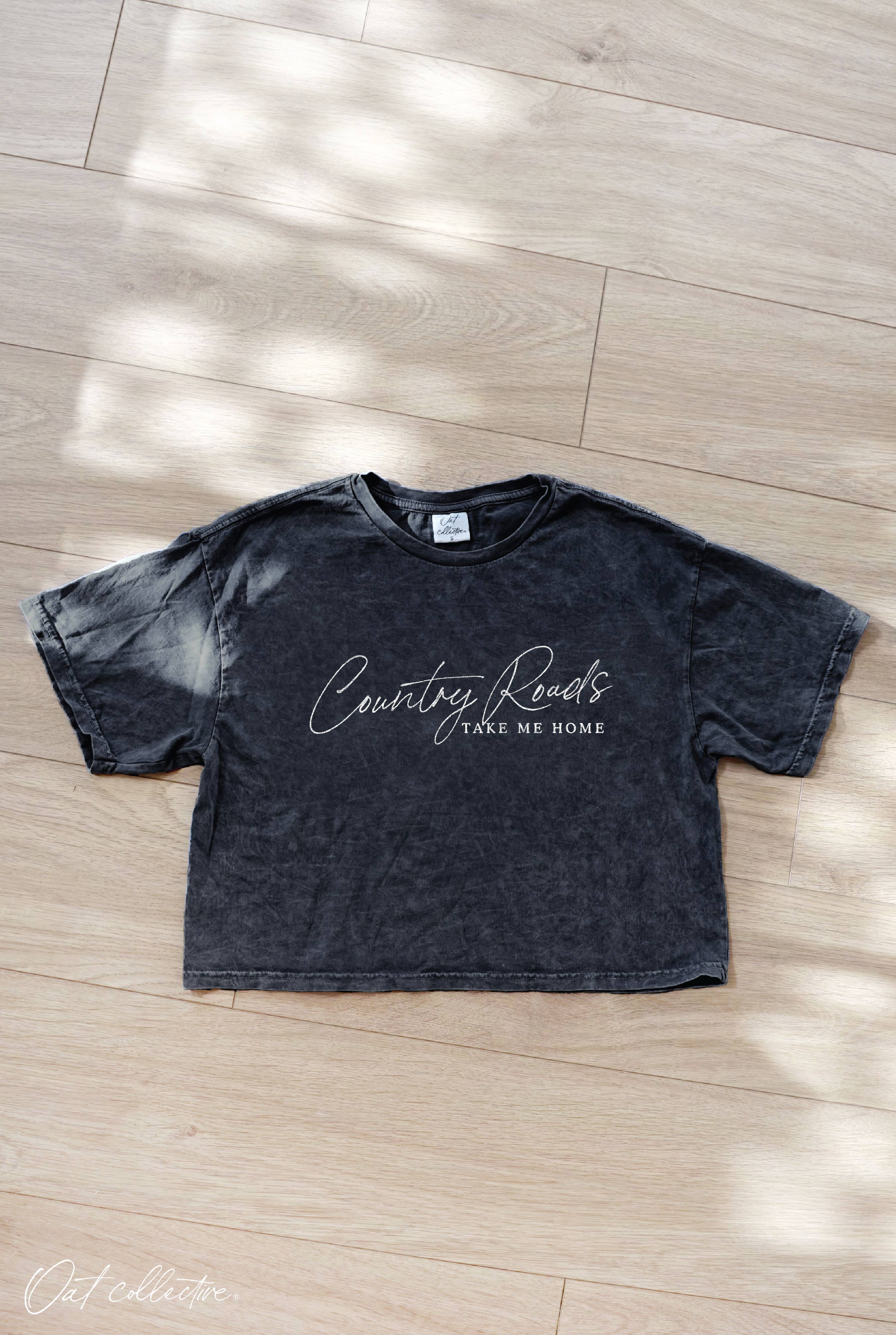 Black t-shirt with 'Country Roads Take Me Home' text on a wooden floor