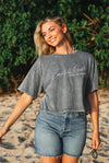 Woman wearing a gray t-shirt with text and denim shorts standing on a beach.