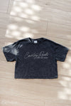 Black t-shirt with 'Country Roads Take Me Home' text on a wooden floor