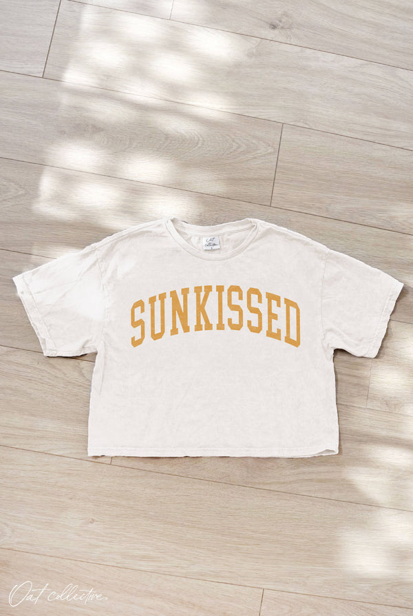 White t-shirt with 'SUNKISSED' text on a wooden floor