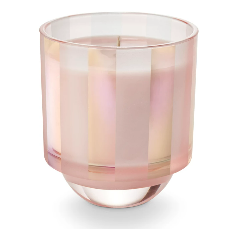 A multicolored striped glass candle, with a pinkish hue at the bottom, sitting inside a box with shiny, tinsel-like details.