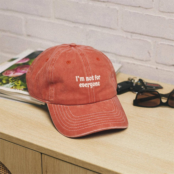 Red cap with 'I'm not for everyone' text on a wooden surface with a magazine and sunglasses.