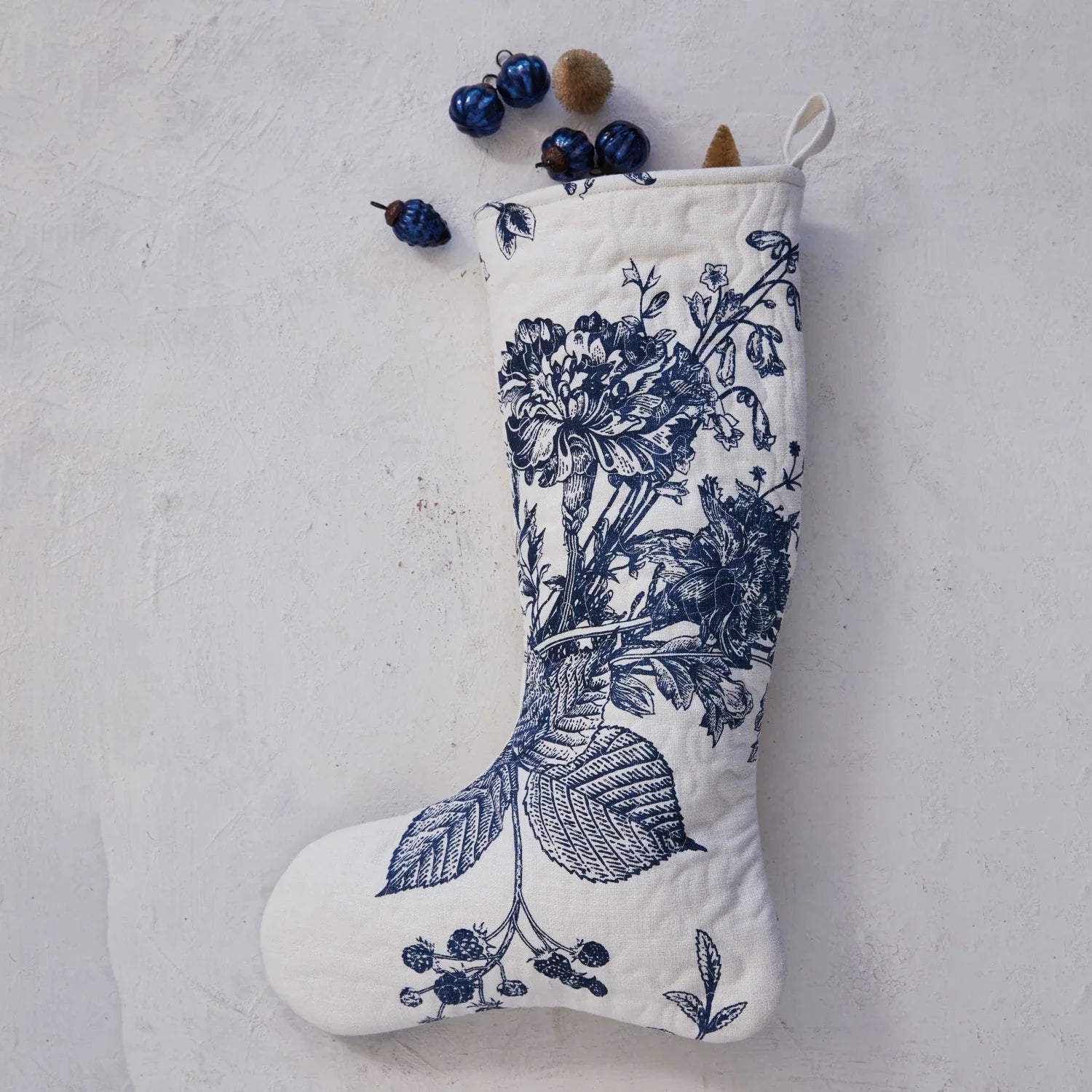 A white Christmas stocking with a blue floral botanical print, displayed against a neutral background with a small ornament for scale.
