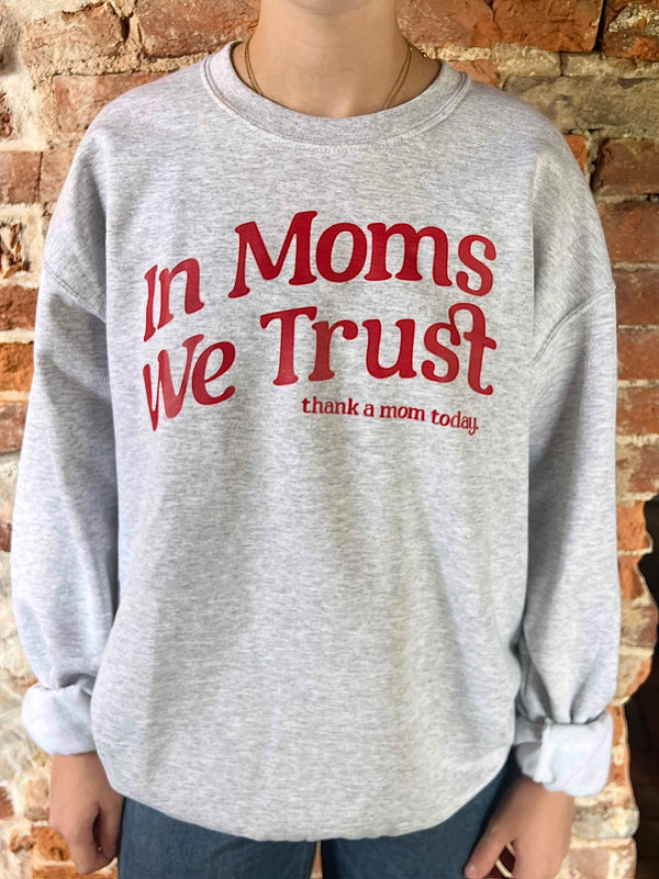 Person wearing a gray sweatshirt with red text 'In Moms We Trust' against a brick wall.