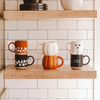 Three ceramic mugs on a wooden shelf against a tiled wall.