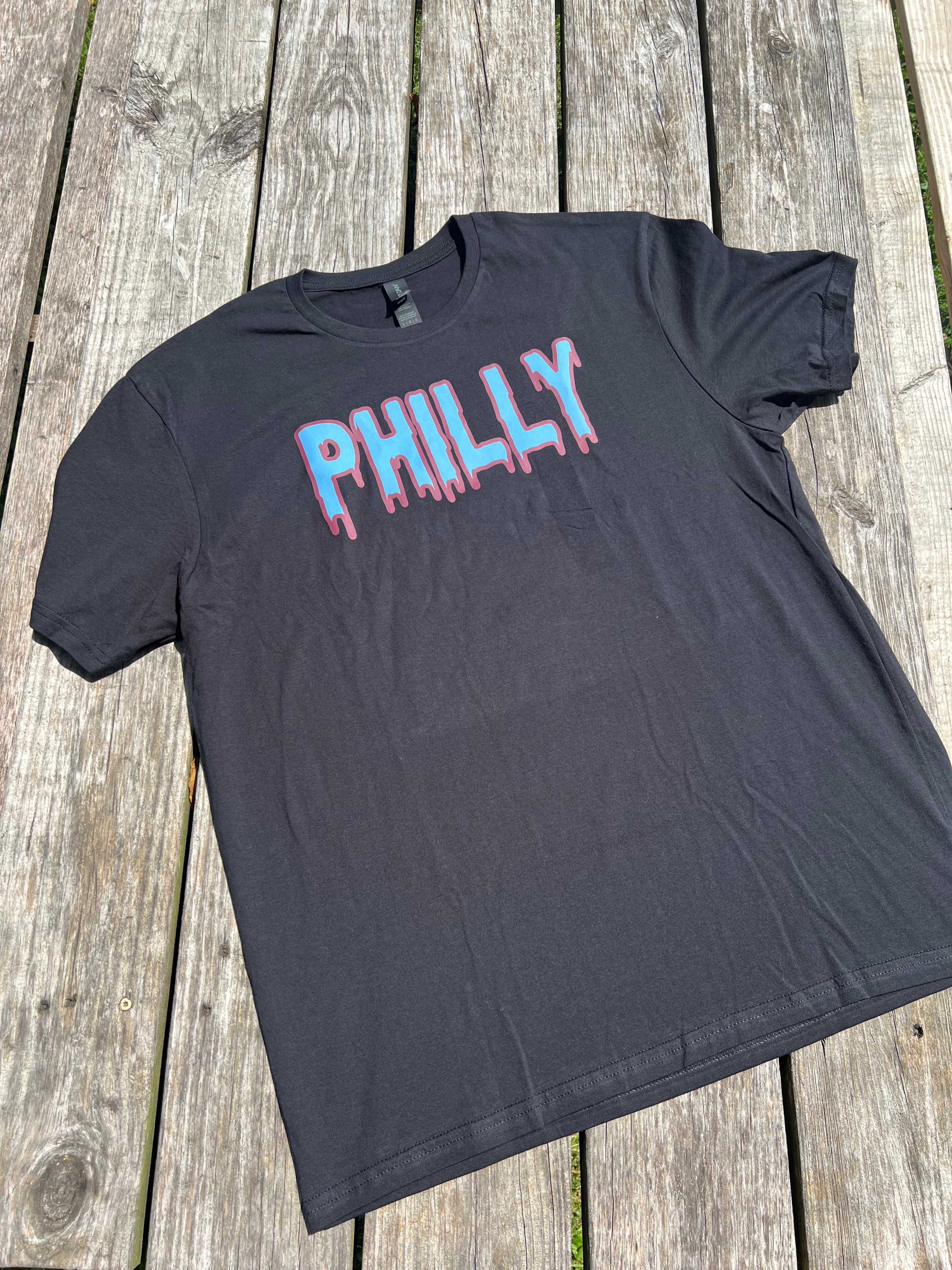 Black t-shirt with 'PHILLY' printed on it in pink and blue, laid flat on a wooden surface.