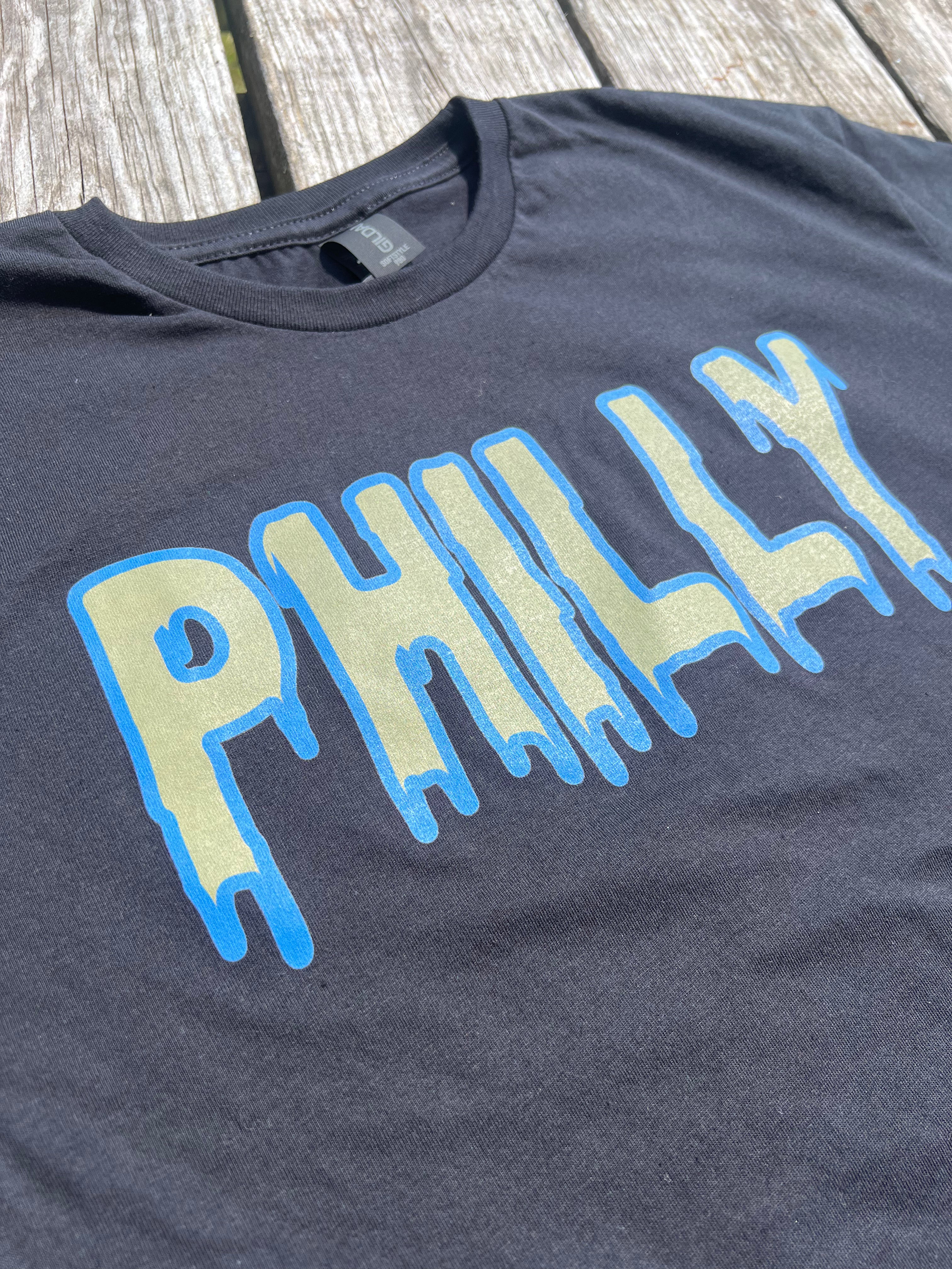 Black t-shirt with 'PHILLY' printed in blue and light green on a wooden surface