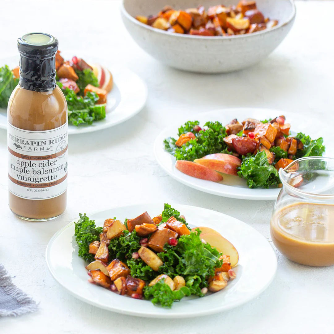 Plates of salad with a bottle of apple cider maple balsamic vinaigrette on a white table.