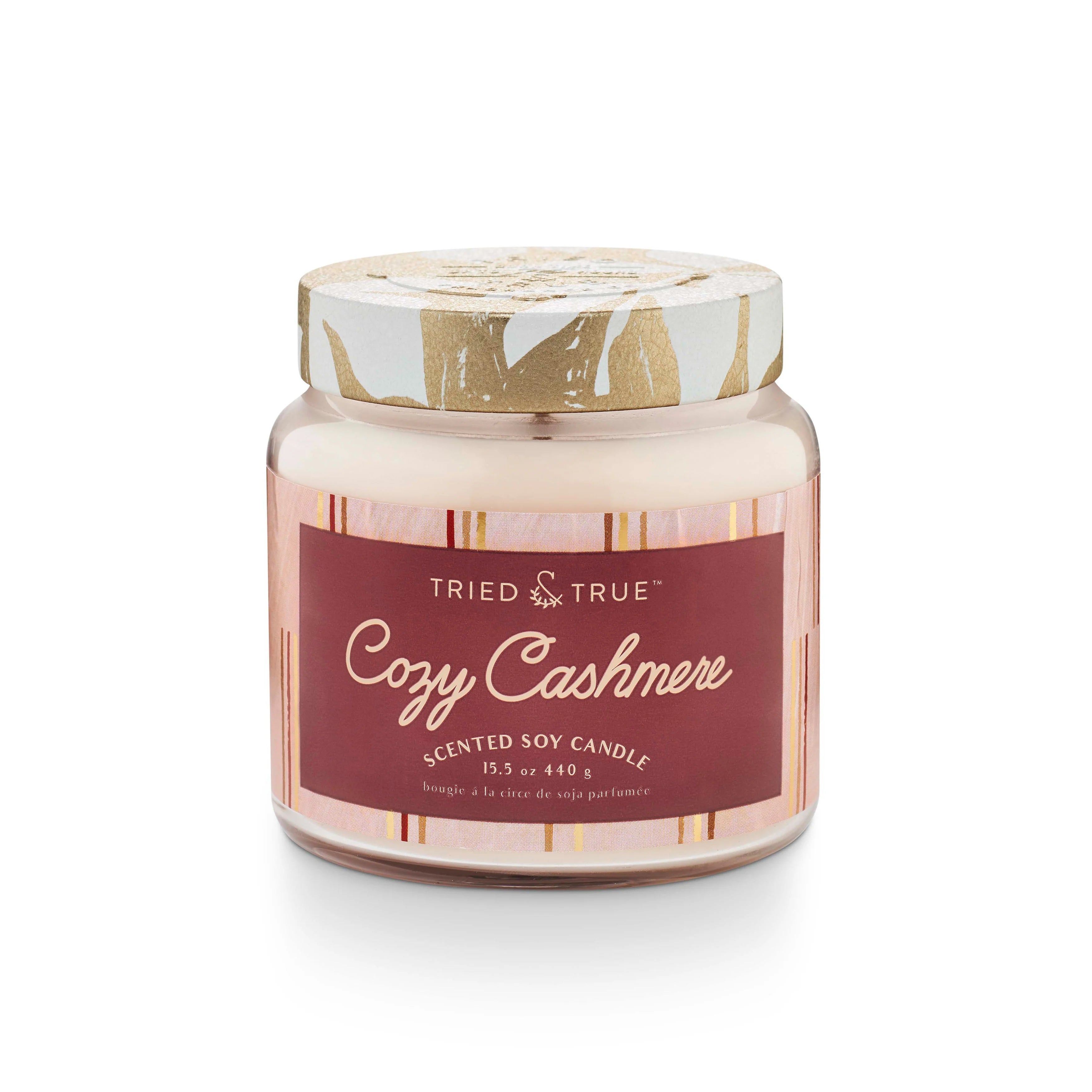 A scented soy candle jar with a label reading 'Cozy Cashmere'. The jar is clear with a white and gold pattern on the lid, and the label has a red background with white text.