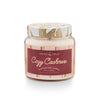 A scented soy candle jar with a label reading 'Cozy Cashmere'. The jar is clear with a white and gold pattern on the lid, and the label has a red background with white text.