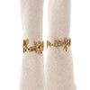 A set of two gold brass napkin rings with the word 'handful of love' written on them, displayed on a textured beige fabric.