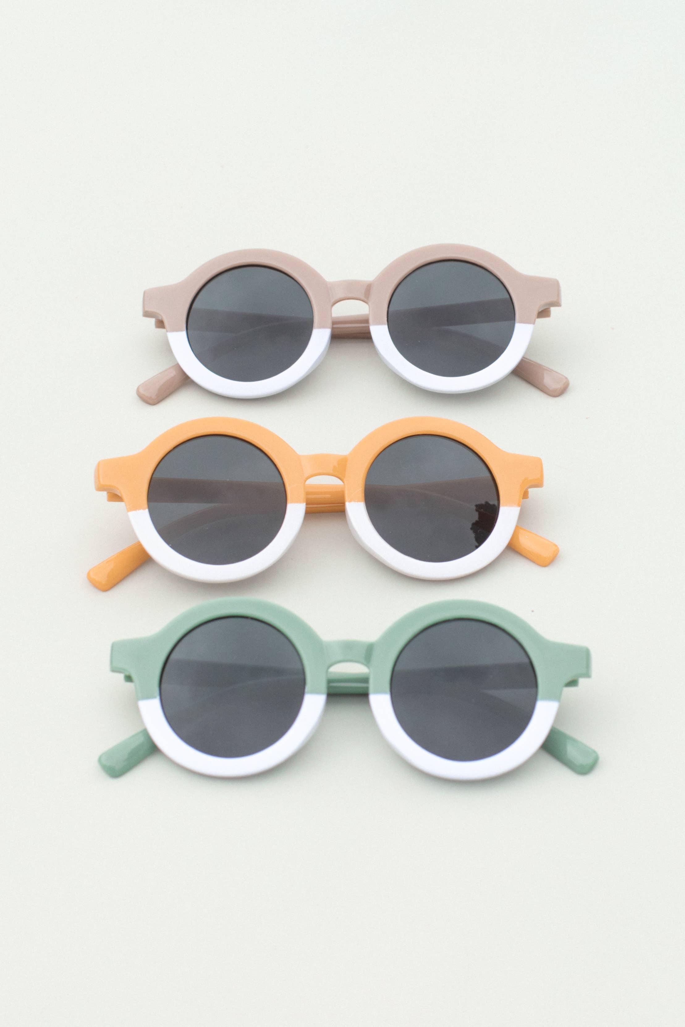 A collection of toddler round sunglasses in three colors: pink, orange, and green, each with white frames and dark lenses.