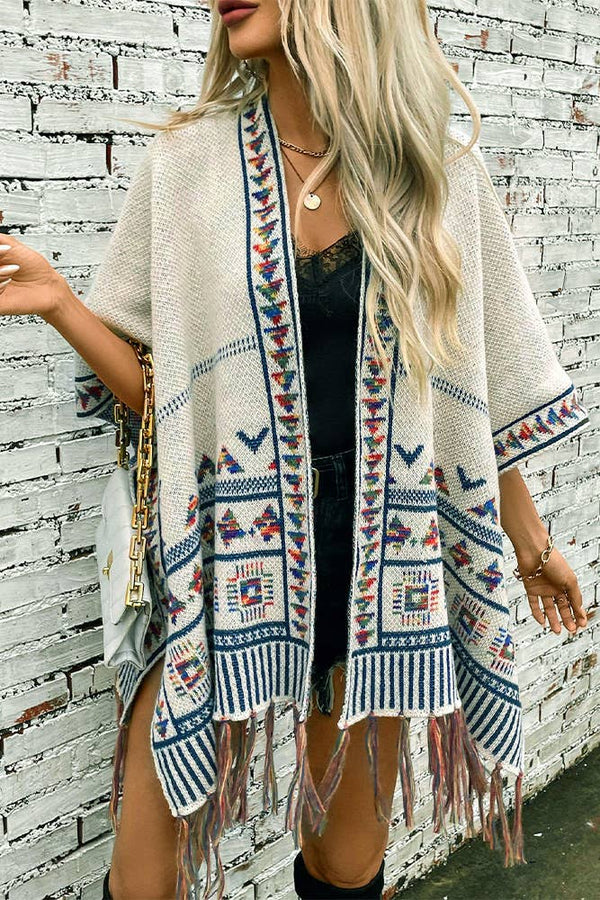 Woman wearing a patterned poncho with fringe against a brick wall.