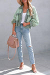 Woman wearing a green jacket, white top, and blue jeans, holding a beige handbag against a gray background.