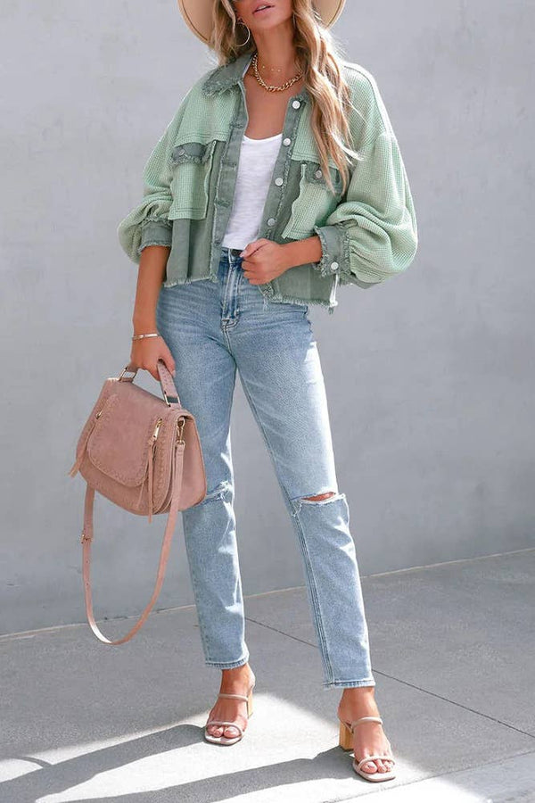 Woman wearing a green jacket, white top, and blue jeans, holding a beige handbag against a gray background.