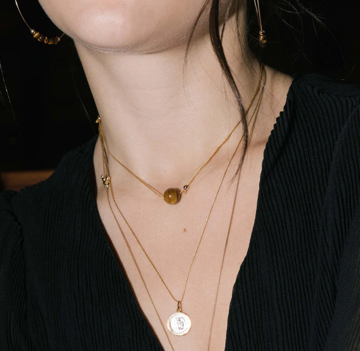 A person wearing a gold-colored adjustable necklace with a single slider bead, featuring a gemstone, and other gold necklaces layered over a shirt.