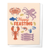 A greeting card with illustrations of various sea creatures such as a crab, fish, and lobster, with the text 'Happy Feasting' displayed at the top.