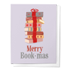 A greeting card with a stack of books wrapped with a red bow and the text 'Merry Book-mas' printed on the front. The card is displayed against a white background.