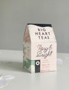 A box of Rosy & Bright tea bags by Big Heart Tea, with a pink and green color scheme, featuring organic rose and hibiscus, cinnamon, and Assam black tea.