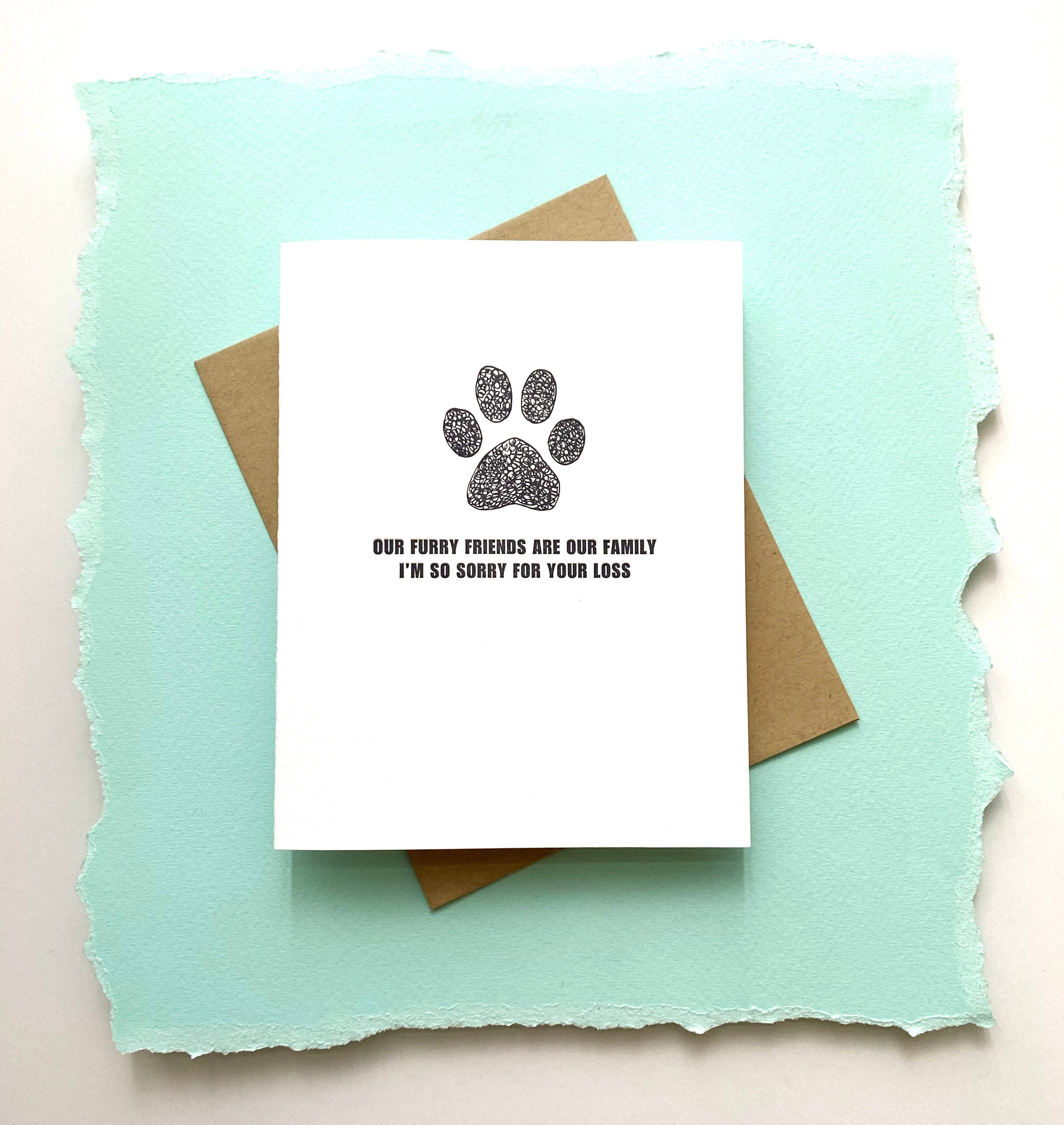 A sympathy card with a white background featuring a black paw print and the text 'Our Furry Friends Are Our Family I'm So Sorry For Your Loss' letterpressed on it, accompanied by a recycled envelope.