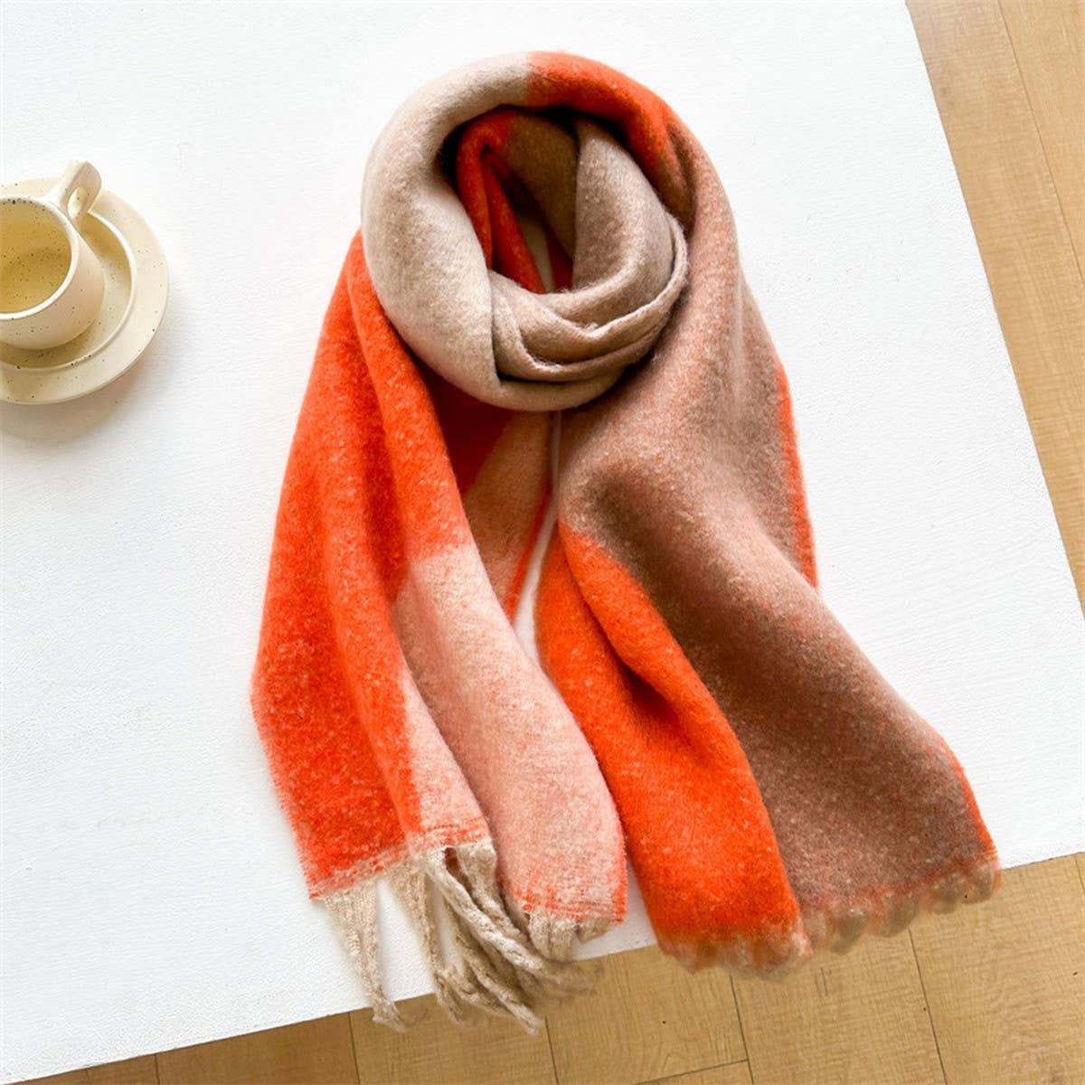 A soft, fuzzy scarf with a color-blocked pattern in orange, beige, and light pink, draped on a white table next to a small cup and saucer.
