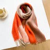 A soft, fuzzy scarf with a color-blocked pattern in orange, beige, and light pink, draped on a white table next to a small cup and saucer.