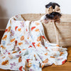A baby swaddle blanket with mushroom illustrations laid out next to a basket with a baby peeking out from inside.