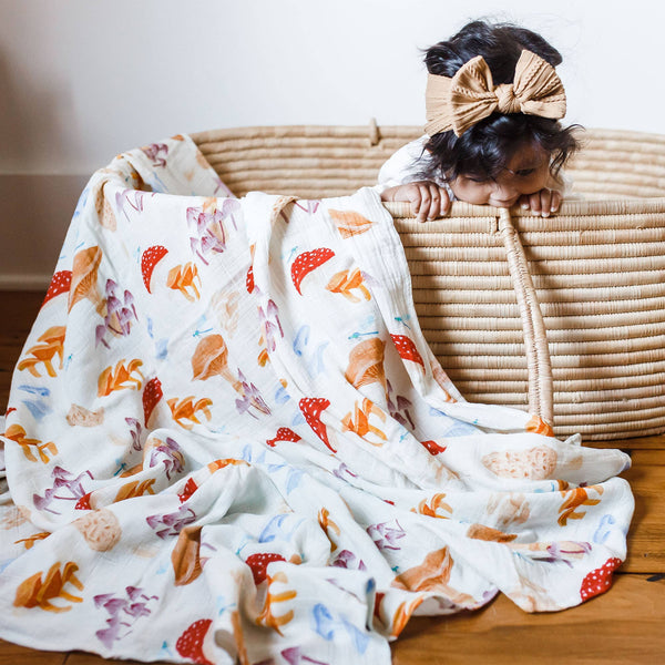 A baby swaddle blanket with mushroom illustrations laid out next to a basket with a baby peeking out from inside.