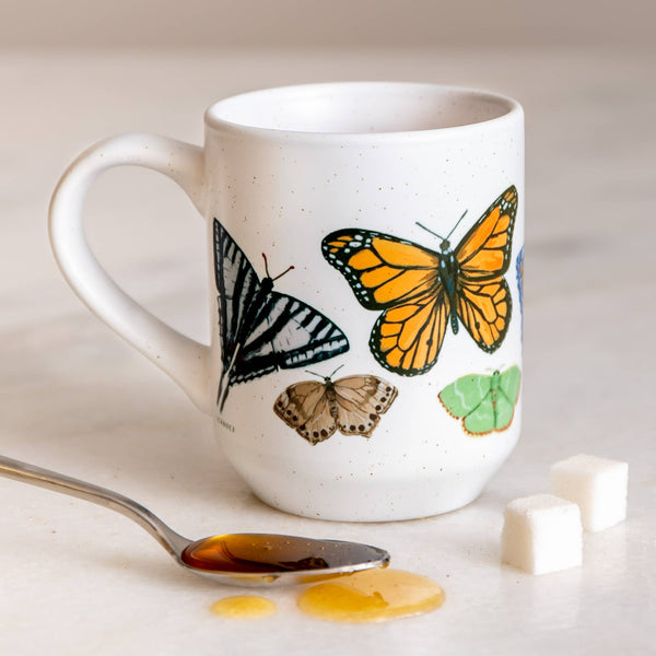 A white ceramic mug with hand-painted illustrations of various butterflies, with a spoon and sugar cubes on a table.