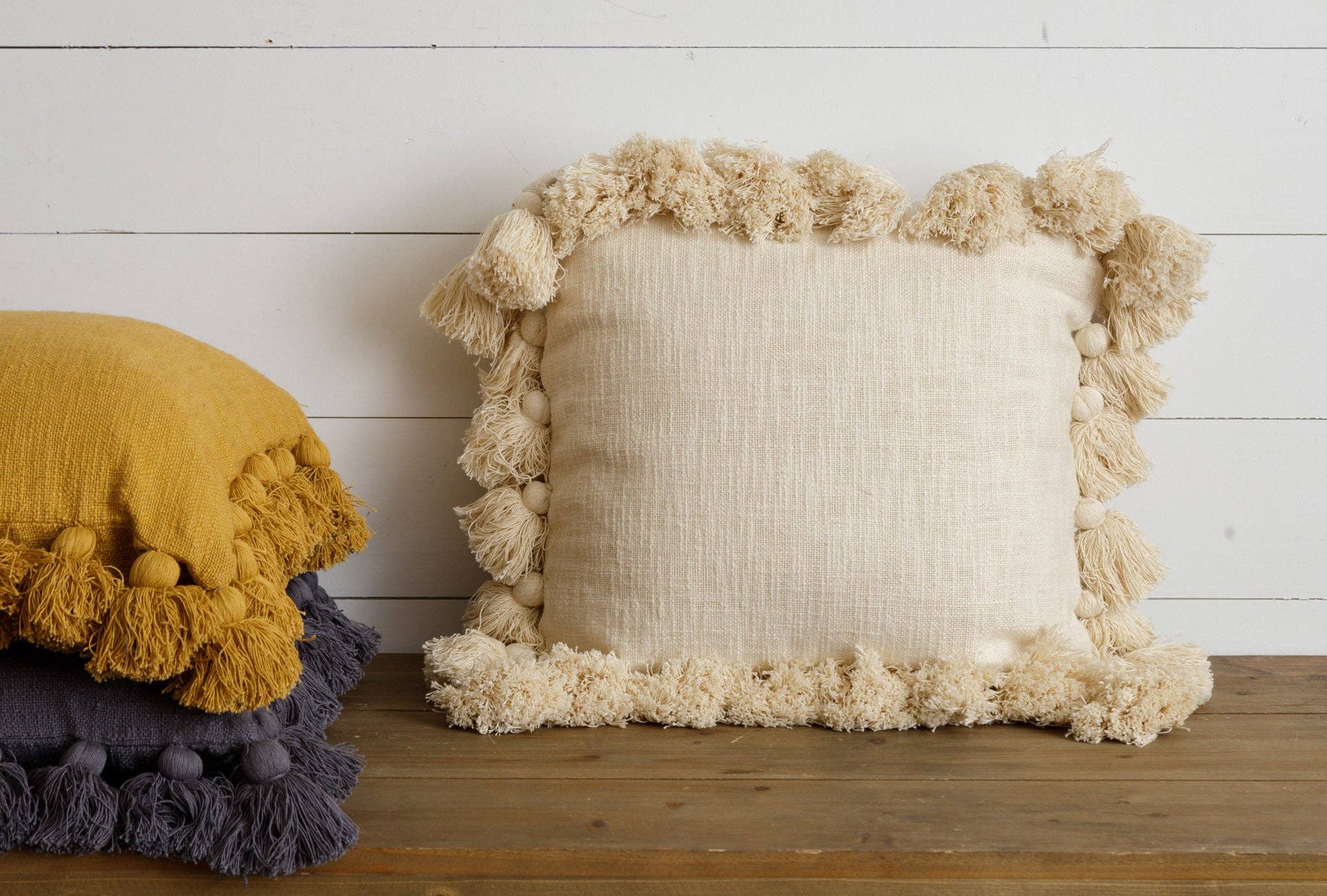 Two square throw pillows, one in yellow and the other in gray, with a tassel design, placed on a wooden surface against a white wall.
