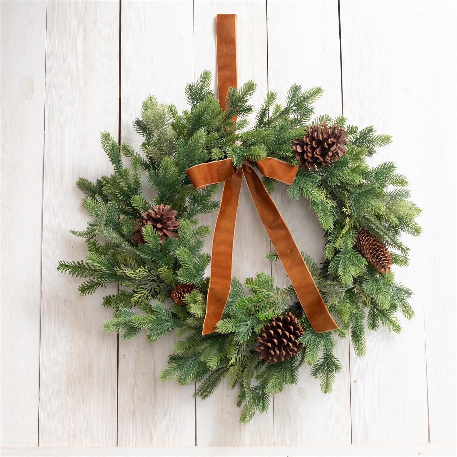 A green pine wreath with brown pinecones and a wide brown ribbon bow, hanging against a white wood plank wall.