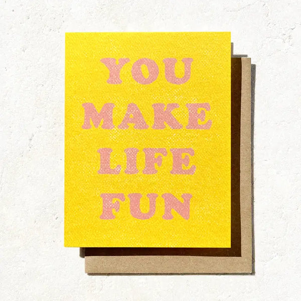 A yellow greeting card with the text 'YOU MAKE LIFE FUN' in pink letters, accompanied by a brown envelope.
