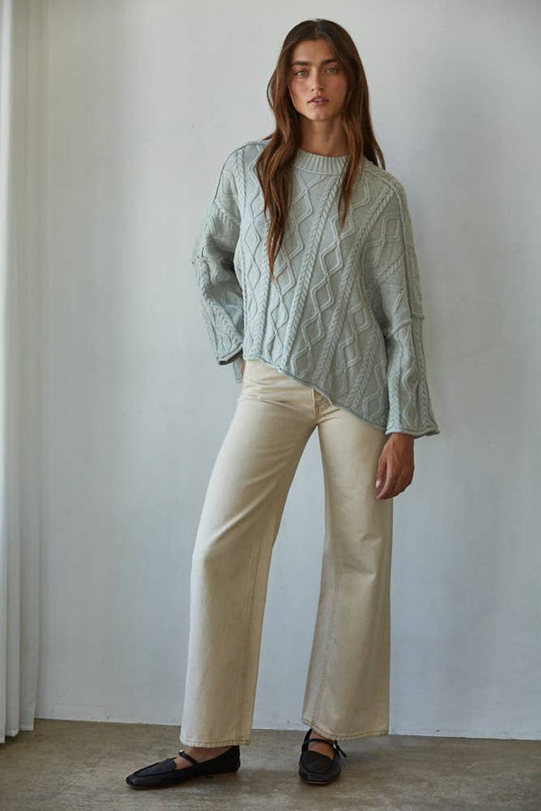 Woman wearing a cable knit sweater and beige pants against a plain background