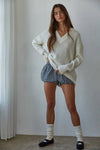 Woman wearing a white sweater and checkered shorts standing against a plain wall.