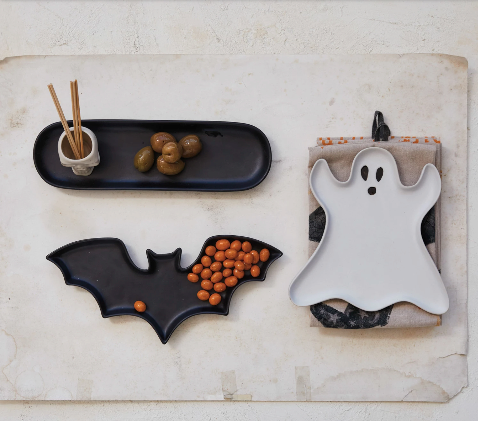 Halloween-themed ceramic plates shaped like a bat, ghost, and black tray on a textured surface.
