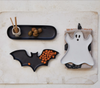 Halloween-themed ceramic plates shaped like a bat, ghost, and black tray on a textured surface.