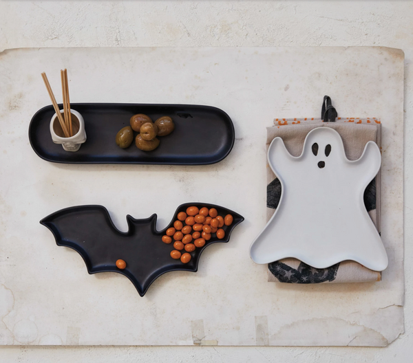 Halloween-themed ceramic plates shaped like a bat, ghost, and black tray on a textured surface.