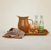 Ceramic pitcher, glass with liquid, and bottles on a tray with a cloth on a light background