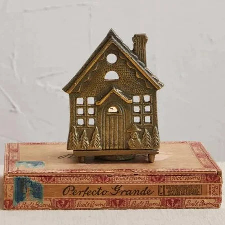 A bronze-colored embossed cast aluminum house tealight holder with open panes and a pointed roof line, placed on top of a cardboard box with 'Perfecto Grande' printed on it.