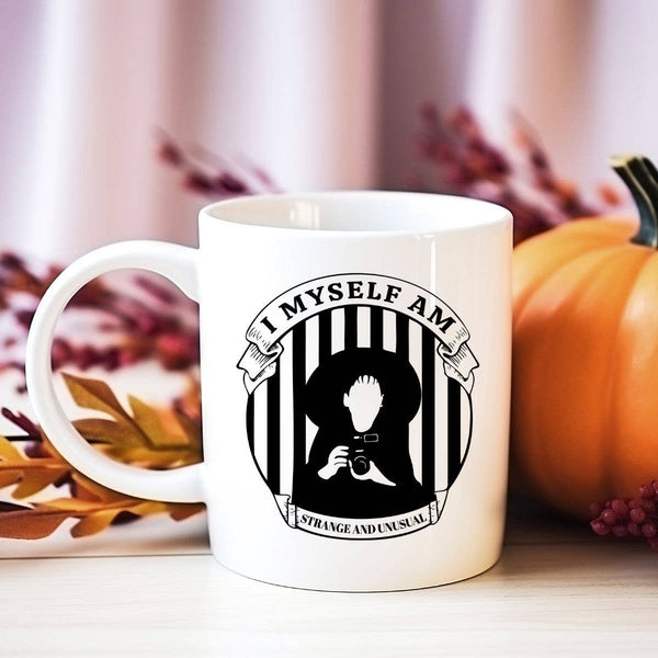 A white coffee mug with a black and white 'I Myself am Strange and Unusual' text and a related graphic, placed on a table with pumpkins and autumn leaves around.