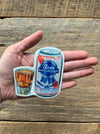 A vinyl sticker designed to look like a can of beer, held in a person's hand against a wooden background.
