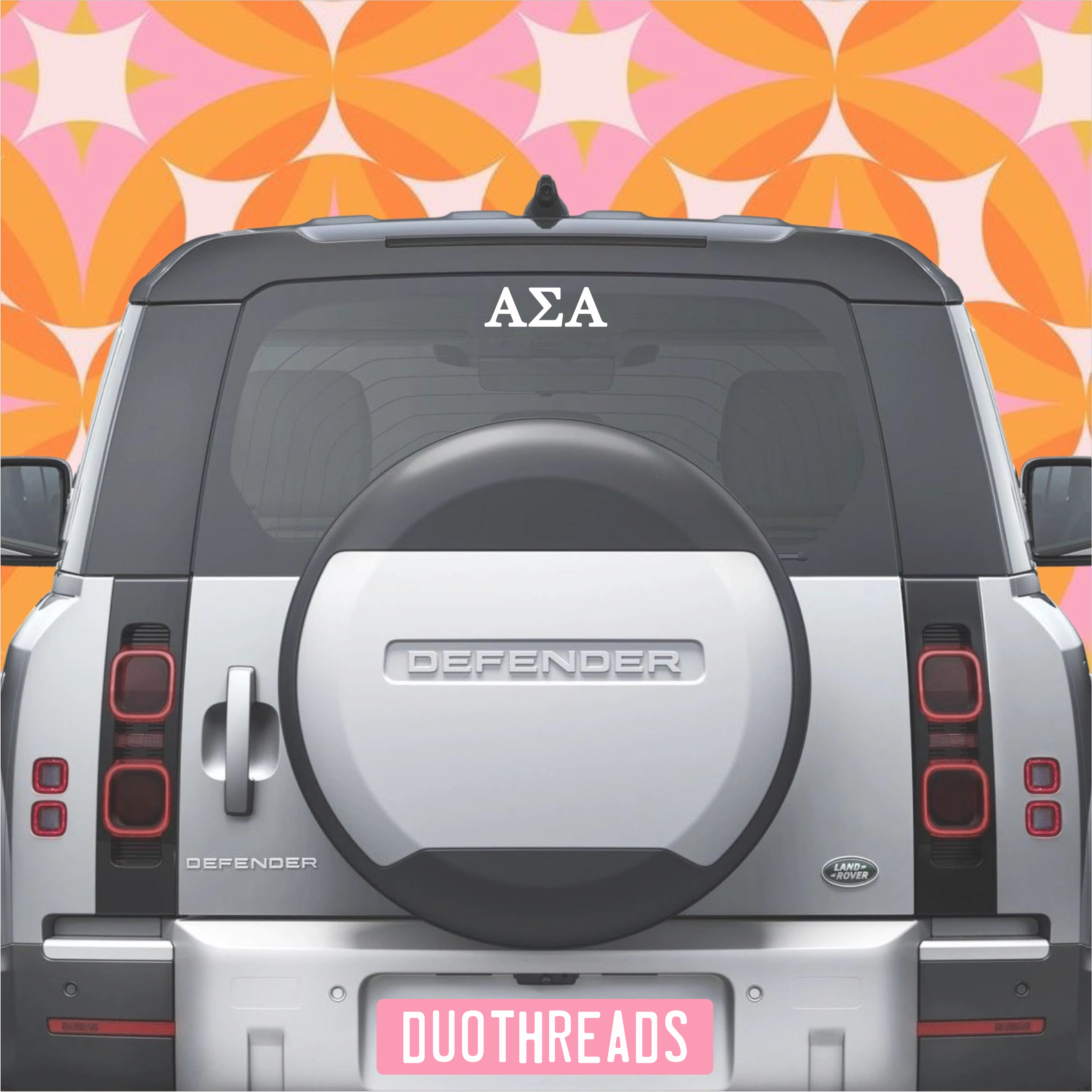 Back of a Land Rover Defender with 'AΣA' on the window against a colorful geometric pattern.