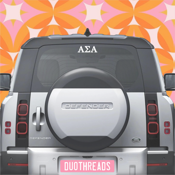 Back of a Land Rover Defender with 'AΣA' on the window against a colorful geometric pattern.