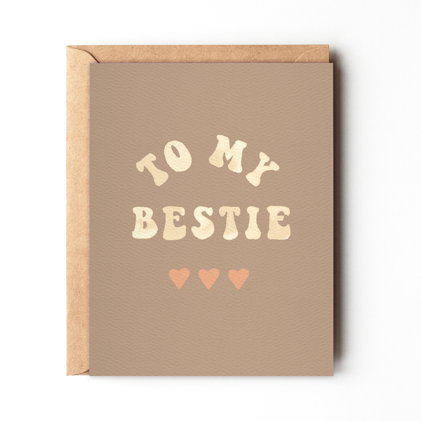 A beige greeting card with the text 'TO MY BESTIE' printed on it, accompanied by two small hearts. A brown envelope is visible behind the card.