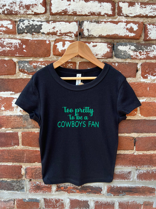 Black t-shirt with 'Too pretty to be a Cowboys fan' text on a wooden hanger against a brick wall.